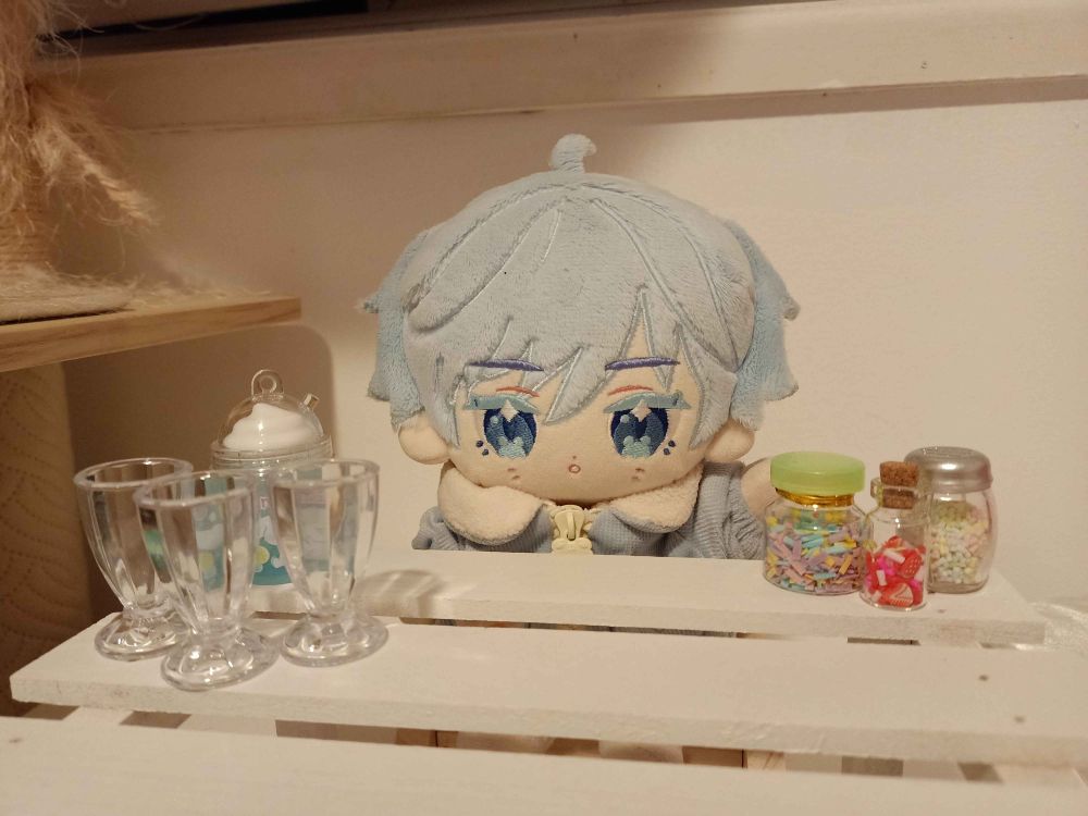 Photo of a Chongyun plushie sitting behind a white wooden table. On the left is some empty milkshake glasses and on the right are some bottles of sprinkles.