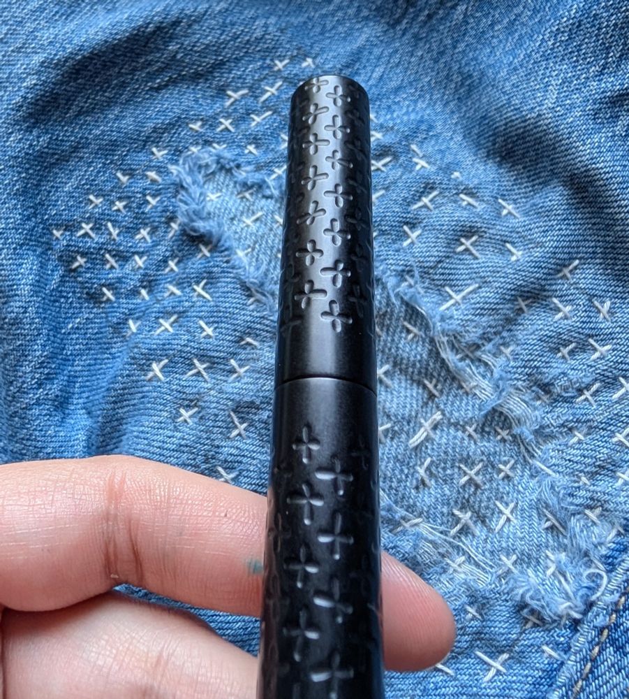 Blue jeans in the background with very uneven white "+" stitches. In the foreground is a black pen with engraved "+" pattern