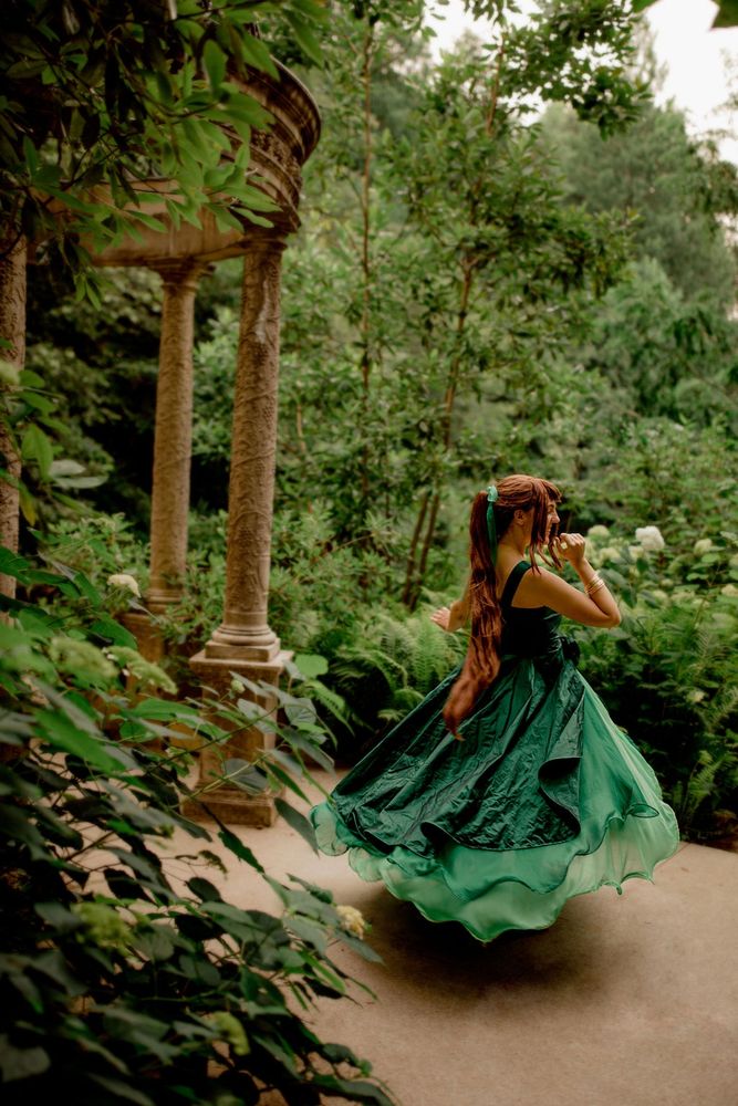 A cosplayer portrays Princess Jupiter from Sailor Moon in her princess dress, which happens to be this year's fan-selected color of the year (Ptalo Green) instead of the officially selected "Cloud Dancer" white. The photo has vivid foliage in the foreground and a gazebo in the midground, where the cosplayer spins and her ruffly skirt flares out dramatically. She's giggling with a hand up near her face. The background is full of additional green foliage.  