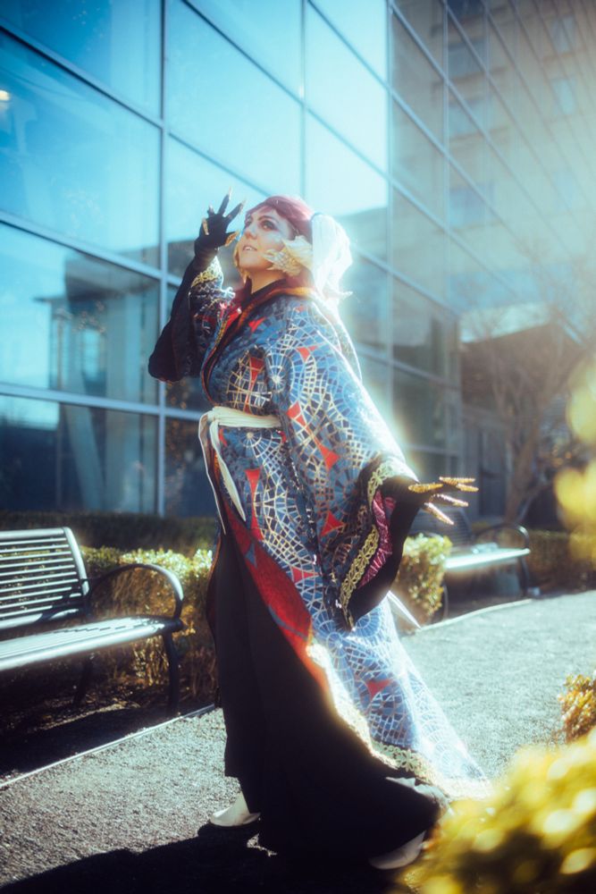 A cosplayer stands in front of a glassy building in a set of wizard robes made from brocade that shares a pattern with the Dragoncon Marriott carpet. She has dragon horns, a tail, and gold claws