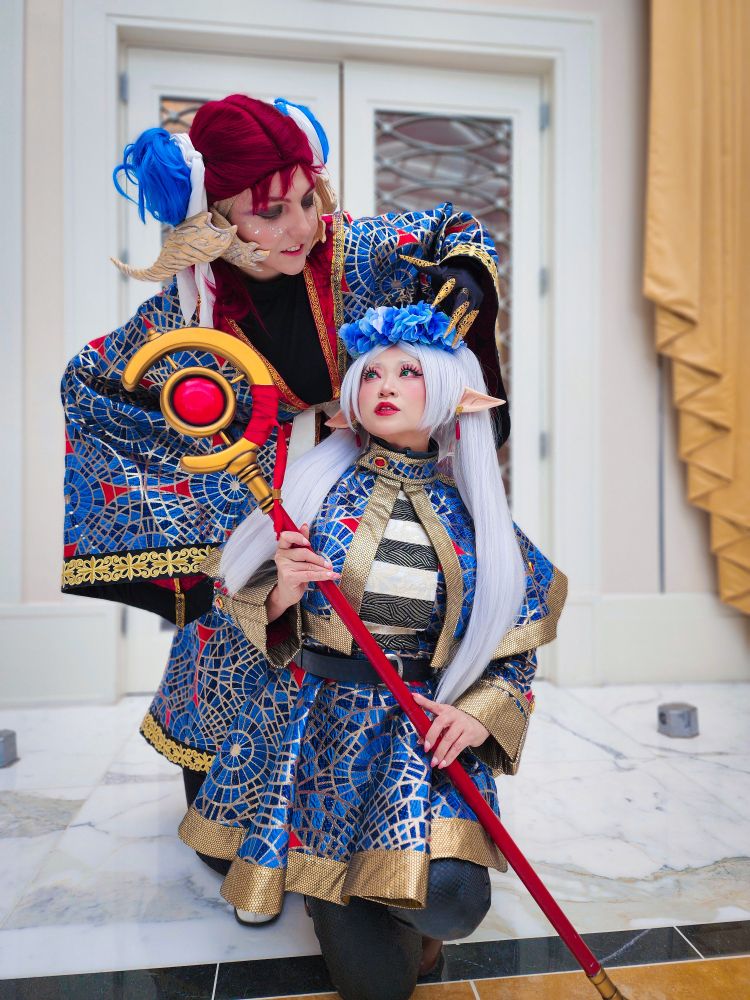 Two cosplayers at Katsucon 2025 wear fantastical outfits made from Yaya Han's Carpet Brocade, a pattern that riffs off of the original Marriott Carpet at Dragoncon. Yaya is wearing her Carpet Frieren, holds her staff, is kneeling on the floor, and is looking up at Cass. Cass is an original character, styled to look like a dragon with horns, golden claws, and a tail, with big wizardy robes in the same fabric. 