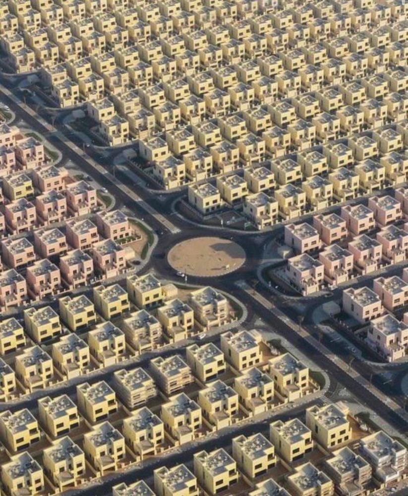 Aerial view of a residential district in Dubai featuring hundreds of identical cube-shaped villas arranged in a precise grid pattern. The homes are painted in pastel tones, including beige, yellow, and pink, and are separated by narrow roads that intersect at regular intervals. A circular roundabout is visible near the center. The highly organized layout emphasizes symmetry and uniformity, characteristic of planned urban developments in the region.