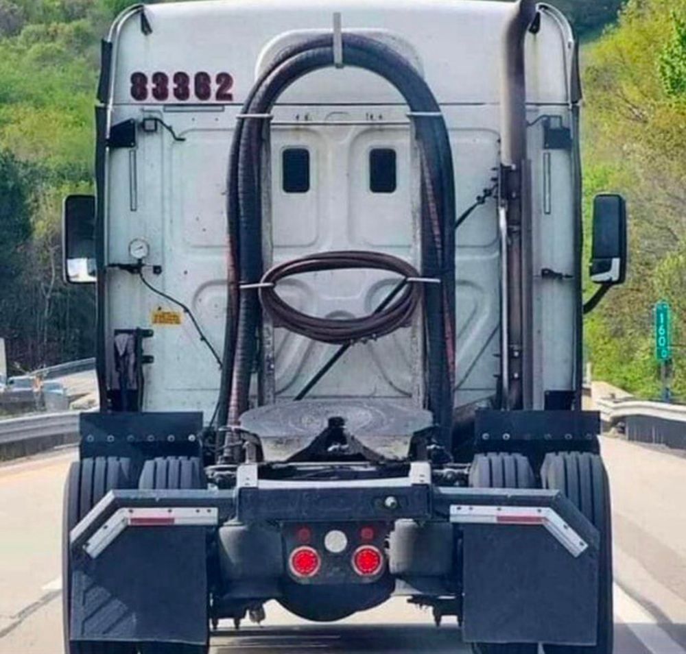 Back of a semi-truck cab seen from the highway. Two large vertical hoses and a coiled cable form the appearance of eyes and hair and a smiling mouth, making the truck look like Jesus
