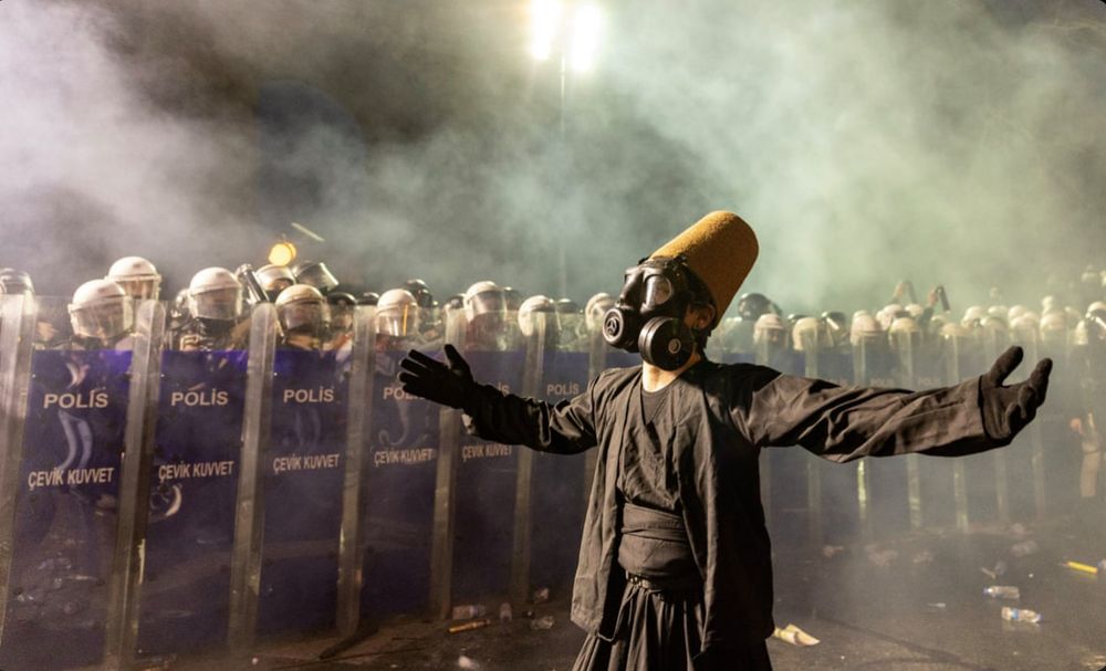 Alt for four images:

	1.	A person in a black robe and a tall brown felt hat stands facing a line of riot police holding shields labeled “Polis.” The person is being sprayed with pepper spray, and the air is filled with mist and debris. The person wears a gas mask and stands with a composed posture amid the chaos.
	2.	The same person in black robes and a tall brown felt hat stands calmly with arms folded while being sprayed with pepper spray by riot police. The individual wears a gas mask and stands on a wet street littered with water bottles. A line of police in helmets and shields labeled “Polis” stands in the background.
	3.	A person wearing a black robe, gas mask, and tall brown felt hat stands with arms outstretched in front of a row of riot police. The police hold transparent shields marked “Polis” and “Çevik Kuvvet.” Smoke fills the air, and the scene is illuminated by strong overhead lights.
	4.	A similar scene shows the same person in a dramatic pose with arms wide open and head tilted back, wearing the same attire. Smoke surrounds the area, and the line of riot police with shields labeled “Polis” and “Çevik Kuvvet” remains in the background. Debris is scattered on the ground.