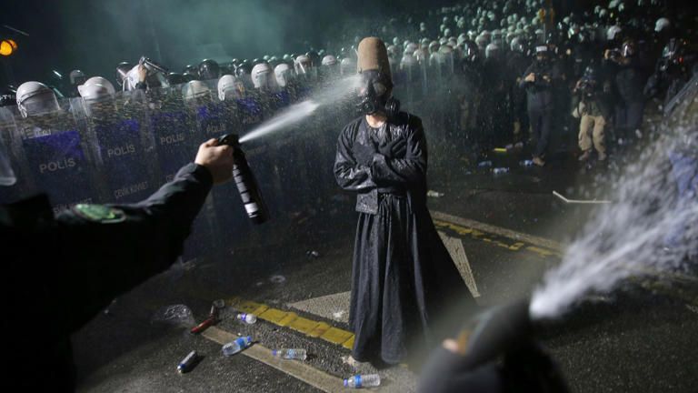 Alt for four images:

	1.	A person in a black robe and a tall brown felt hat stands facing a line of riot police holding shields labeled “Polis.” The person is being sprayed with pepper spray, and the air is filled with mist and debris. The person wears a gas mask and stands with a composed posture amid the chaos.
	2.	The same person in black robes and a tall brown felt hat stands calmly with arms folded while being sprayed with pepper spray by riot police. The individual wears a gas mask and stands on a wet street littered with water bottles. A line of police in helmets and shields labeled “Polis” stands in the background.
	3.	A person wearing a black robe, gas mask, and tall brown felt hat stands with arms outstretched in front of a row of riot police. The police hold transparent shields marked “Polis” and “Çevik Kuvvet.” Smoke fills the air, and the scene is illuminated by strong overhead lights.
	4.	A similar scene shows the same person in a dramatic pose with arms wide open and head tilted back, wearing the same attire. Smoke surrounds the area, and the line of riot police with shields labeled “Polis” and “Çevik Kuvvet” remains in the background. Debris is scattered on the ground.