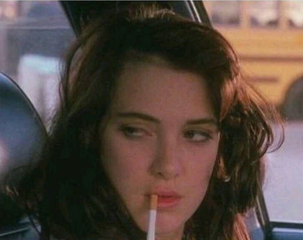 A young woman with shoulder-length dark brown hair sits in a car, holding an unlit cigarette between her lips. She looks to her right with a fuck this expression. A yellow school bus is visible through the window in the background.