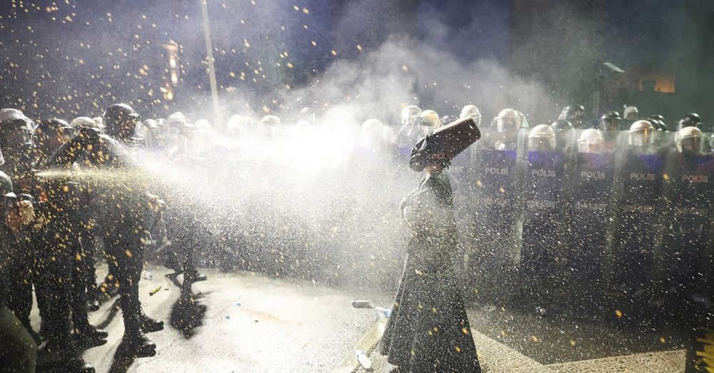 Alt for four images:

	1.	A person in a black robe and a tall brown felt hat stands facing a line of riot police holding shields labeled “Polis.” The person is being sprayed with pepper spray, and the air is filled with mist and debris. The person wears a gas mask and stands with a composed posture amid the chaos.
	2.	The same person in black robes and a tall brown felt hat stands calmly with arms folded while being sprayed with pepper spray by riot police. The individual wears a gas mask and stands on a wet street littered with water bottles. A line of police in helmets and shields labeled “Polis” stands in the background.
	3.	A person wearing a black robe, gas mask, and tall brown felt hat stands with arms outstretched in front of a row of riot police. The police hold transparent shields marked “Polis” and “Çevik Kuvvet.” Smoke fills the air, and the scene is illuminated by strong overhead lights.
	4.	A similar scene shows the same person in a dramatic pose with arms wide open and head tilted back, wearing the same attire. Smoke surrounds the area, and the line of riot police with shields labeled “Polis” and “Çevik Kuvvet” remains in the background. Debris is scattered on the ground.
