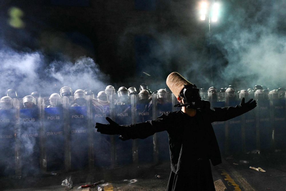 Alt for four images:

	1.	A person in a black robe and a tall brown felt hat stands facing a line of riot police holding shields labeled “Polis.” The person is being sprayed with pepper spray, and the air is filled with mist and debris. The person wears a gas mask and stands with a composed posture amid the chaos.
	2.	The same person in black robes and a tall brown felt hat stands calmly with arms folded while being sprayed with pepper spray by riot police. The individual wears a gas mask and stands on a wet street littered with water bottles. A line of police in helmets and shields labeled “Polis” stands in the background.
	3.	A person wearing a black robe, gas mask, and tall brown felt hat stands with arms outstretched in front of a row of riot police. The police hold transparent shields marked “Polis” and “Çevik Kuvvet.” Smoke fills the air, and the scene is illuminated by strong overhead lights.
	4.	A similar scene shows the same person in a dramatic pose with arms wide open and head tilted back, wearing the same attire. Smoke surrounds the area, and the line of riot police with shields labeled “Polis” and “Çevik Kuvvet” remains in the background. Debris is scattered on the ground.