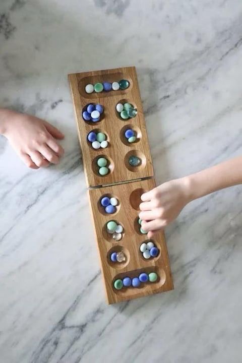 A wooden Mancala board with small round pits filled with glass beads in shades of blue, green, and white is placed on a marble surface. The board is open, showing two rows of pits and larger storage wells at each end. Two children’s hands are visible; one hand is reaching to move beads, while the other rests nearby.