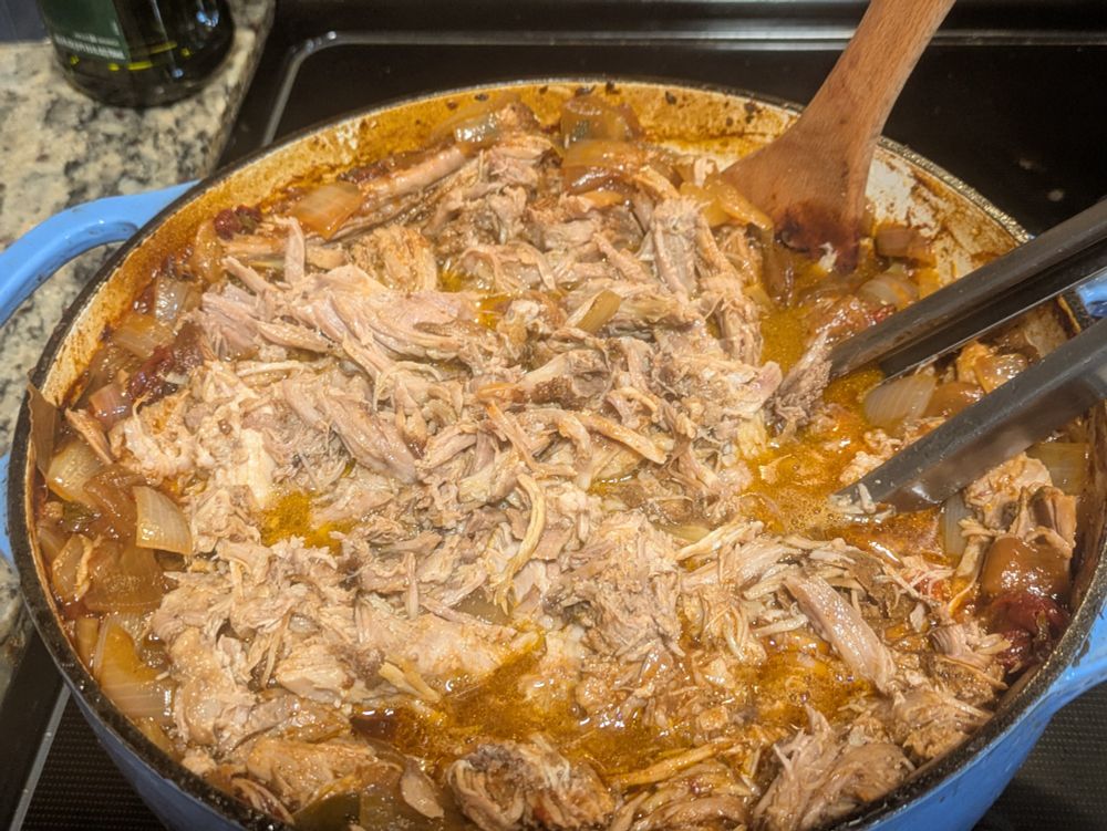 An 8qt Dutch oven filled to the brim with pulled pork in red chili sauce.