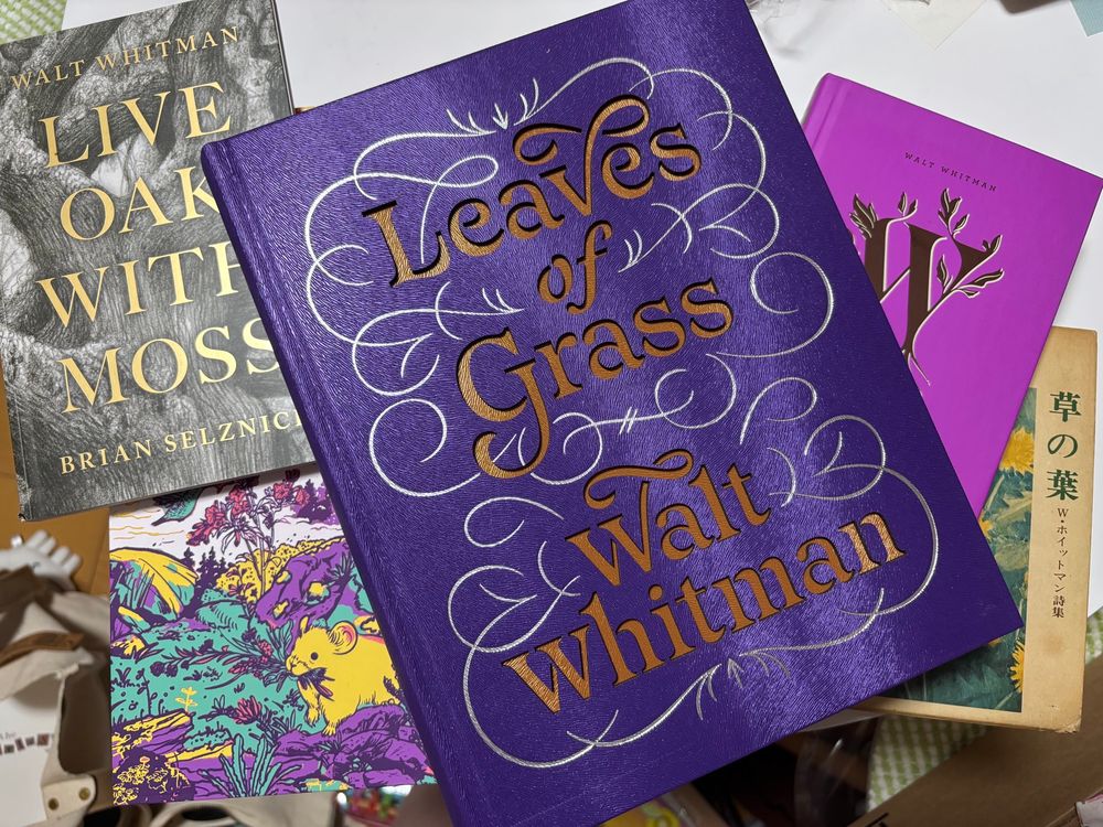 Photo of a stack of books. In the middle is a large purple book with inlaid print and swirling decoration in gold and white that says “Leaves of Grass Walt Whitman”. On the right you can see another brighter purple book with a large W on the cover, decorated with leaves. To the right of that is an older-looking Japanese book with an image of dandelions and the title 草の葉. On the left of the large purple book is a grey book with the detail of a tree and “Live Oak With Moss” on the title. Below that is a brightly colored slipcase showing art of a small rodent eating some grass. 