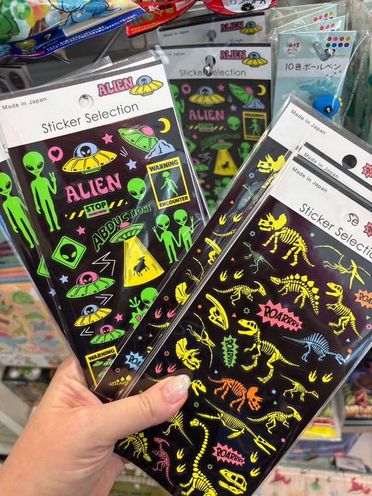 Photo of two sheets of stickers with black backgrounds and neon colors showing dinosaur skeletons and aliens with text like “ROAR” and “ABDUCTION!”