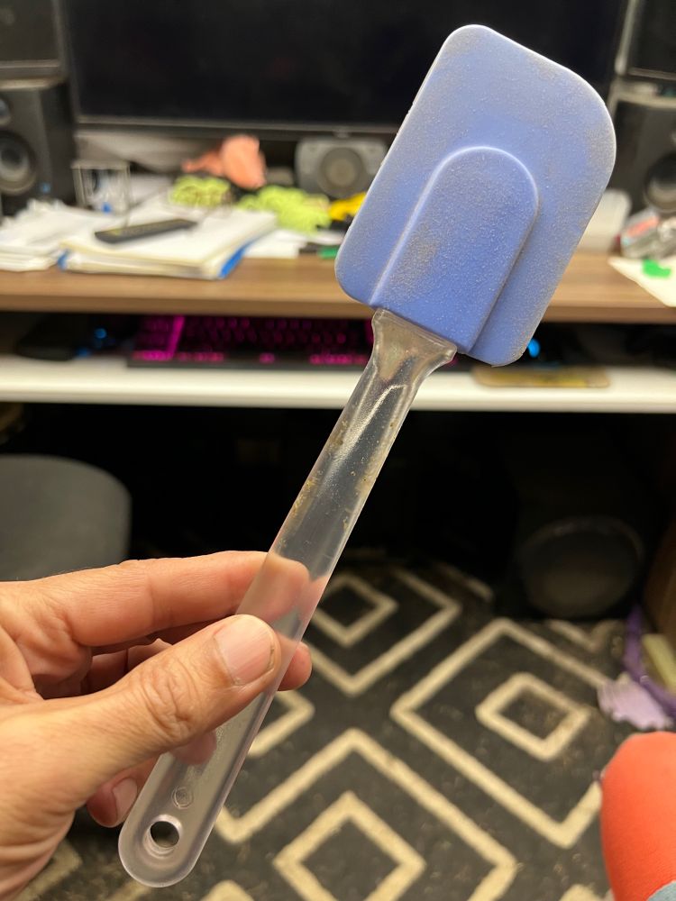 A blue silicone spatula with a clear handle.
Being held by a hand in front of camera.
Background is carpet and a desk and computer keyboard.