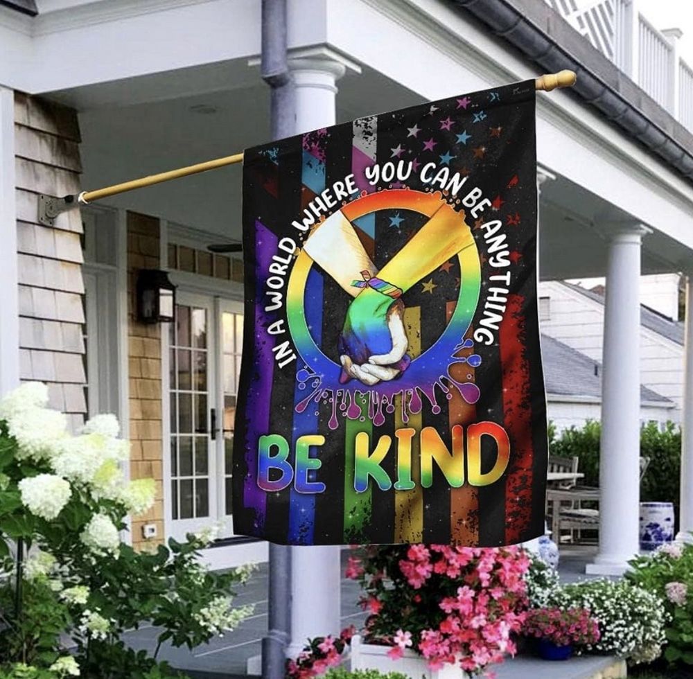 On a cloudy day, a United States flag hangs from a pole in front of a white portico with columns surrounded by flowers in their beds. The flag is somewhat covered by various rainbow colors and includes a circle, within which are two arms, hands entwined, forming a peace symbol, and around its perimeter is written: “In a world where you can be anything, be kind.”