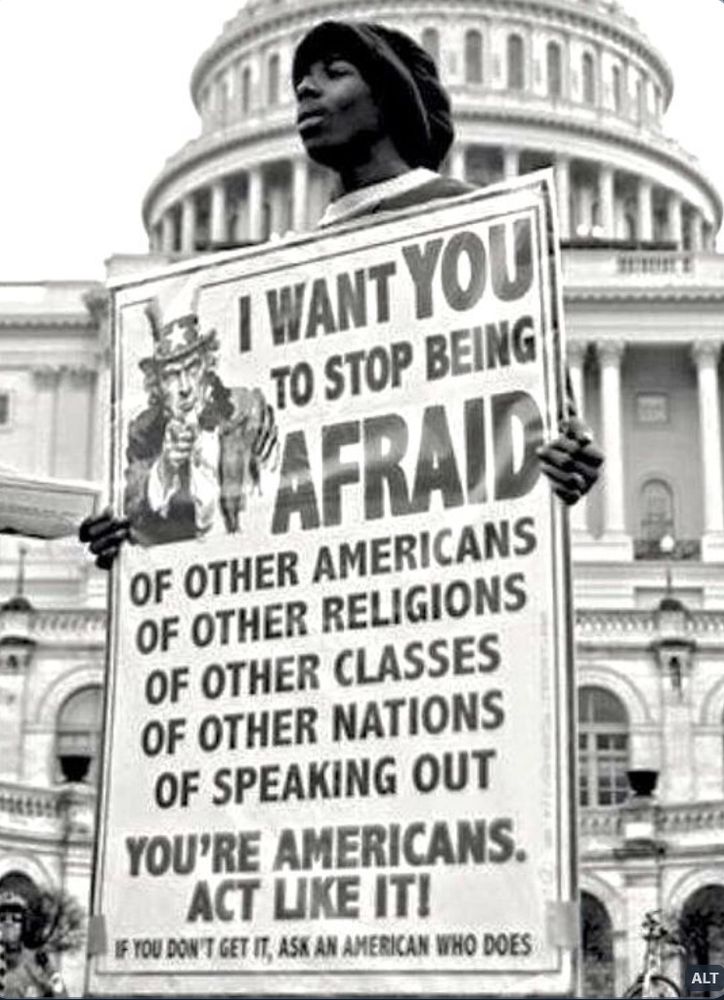 A black and white photo of a young black man, wearing a cap, standing outside of the U.S. Capitol on an overcast day, and holding a large sign that depicts ‘Uncle Sam' wearing his one-starred top hat and pointing to the viewer, that reads: “I want you to stop being afraid of other Americans, of other religions, of other classes, of other nations, of speaking out. You’re Americans. Act like it! If you don’t get it, ask an American who does.” 