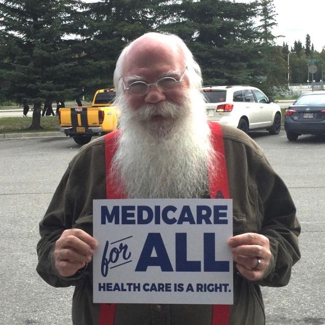 Santa Claus is wearing his wire-rim glasses, green shirt and red suspenders and standing outside, on a clear day in a parking lot with green trees in the background, and holding a sign that reads: “Medicare for All - Health Care is a Right.”