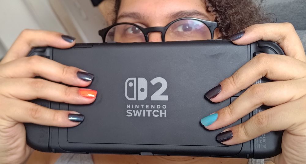 Foto cortada de perto das costas de um switch 2 segurado por uma mulher negra de pele clara, o switch cobre o rosto dela mostrando só os olhos, óculos e parte do cabelo cacheado. As mãos delas estão com as unhas pintadas de preto, exceto pelos anelares, em uma mão a unha anelar está vermelho neon e a outra azul neon, imitando o esquema de cor do switch
