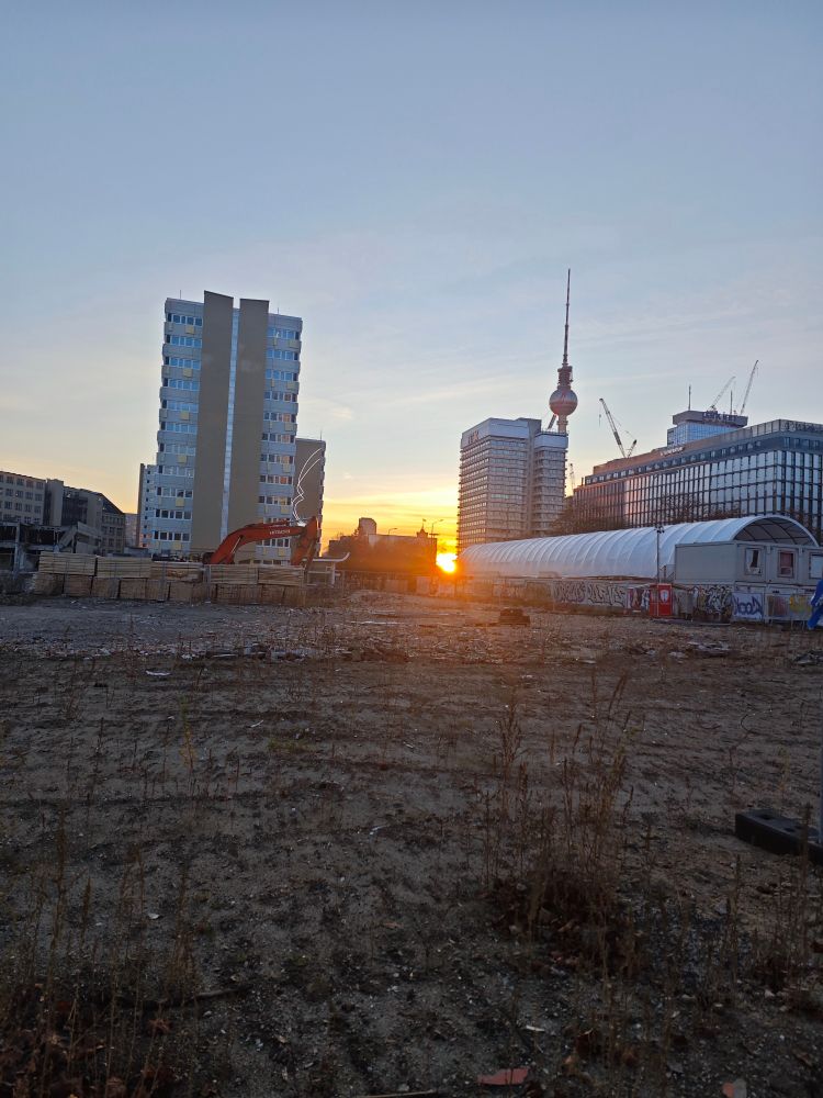 Im Vordergrund eine brach liegende Fläche mit Bauschuttresten. Im Hintergrund diverse Gebäude samt Berliner Fernsehturm. Zwischen den Gebäuden geht die Sonne unter.