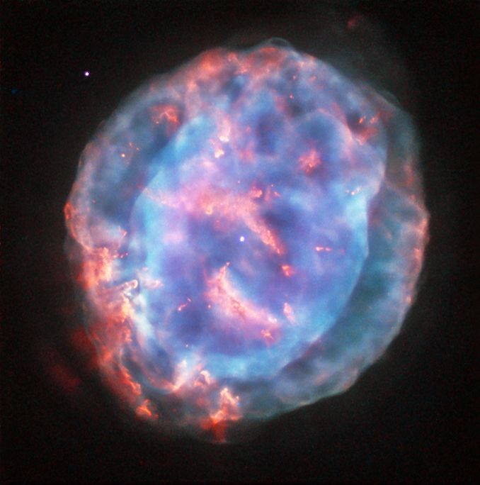 A large, semi-transparent pink, purple, and blue circular object with irregular edges and layers is set against the black background of space. At the center is a very small white star that looks like a dot. The irregular circle, known as the Little Gem Nebula, takes up most of the view. Thicker pink regions appear as arcs above and below the central star, and along the edges. These pink regions have finger-like protrusions and knots of denser gas, which point to the central star. More pink is between 9 o’clock and 6 clock, and near 12 o’clock. There is a little pink near 3 o’clock. There is a semi-translucent purple area in the center. A blue area extends more clearly beyond the purple, running to the edges except where the pink is most prominent. There is one clear white dot toward the top left.