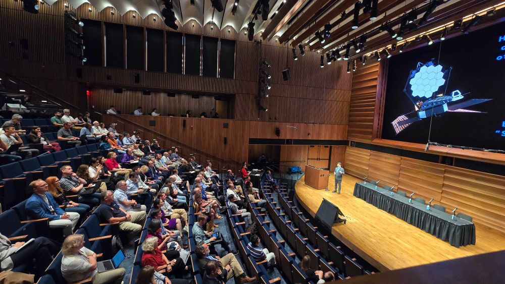 Audience seated in an amphitheater-style lecture hall, attending a presentation about space exploration, featuring visuals of a spacecraft on a large screen.