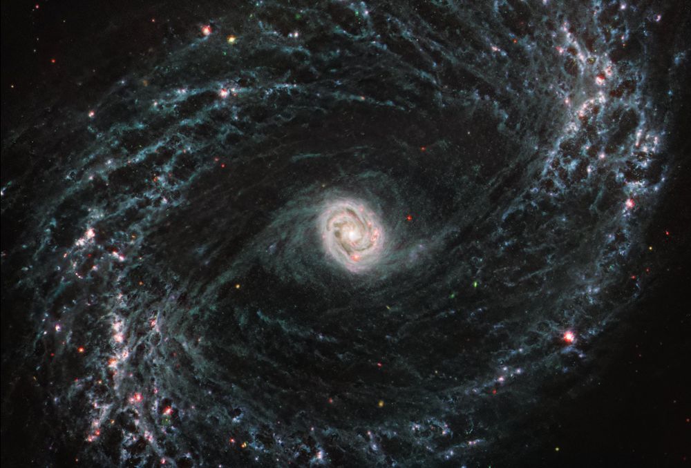 A large, face-on spiral galaxy takes up the entirety of the frame. The scene is mostly black with a bright, glowing oval core at the center. The core is mostly bright white, but there are also swirling, detailed structures that resemble water circling a drain. There is white- and light-blue-colored dust that emanates from the core’s center, but it is tightly contained to the core. There is black space between the core and glowing dust of the outer oval rings. The rings are wispy and highlight filaments of dust around cavernous black bubbles. The dust in the outer rings contains dots that are navy blue, pinkish, reddish, and white. Throughout, there is also a smattering of background galaxies seen as small red and greenish dots.