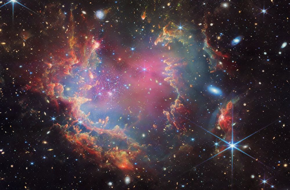 A star cluster shown inside a large nebula that has many colors of gas and dust. The material forms a circle of dark ridges and peaks surrounding the cluster, lit on the inner side, while layers of diffuse, translucent clouds blanket over them. Around and within the gas, a huge number of distant galaxies can be seen, some quite large, as well as a few stars nearer to us which are very large and bright. In the corners, the background of space is black.