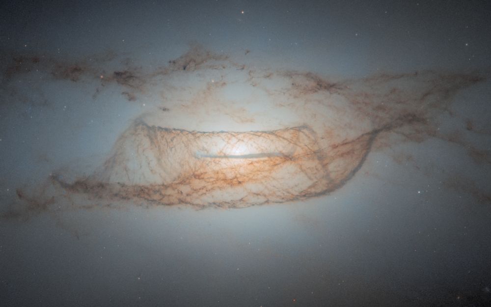 The central portion of a lenticular galaxy. The center holds a bright-white core that illuminates most of the image. The brightness decreases with distance from the center. Water-like waves of dark brown dust surround the center with long clouds of brown dust slightly above. Faint red stars are spread throughout. 