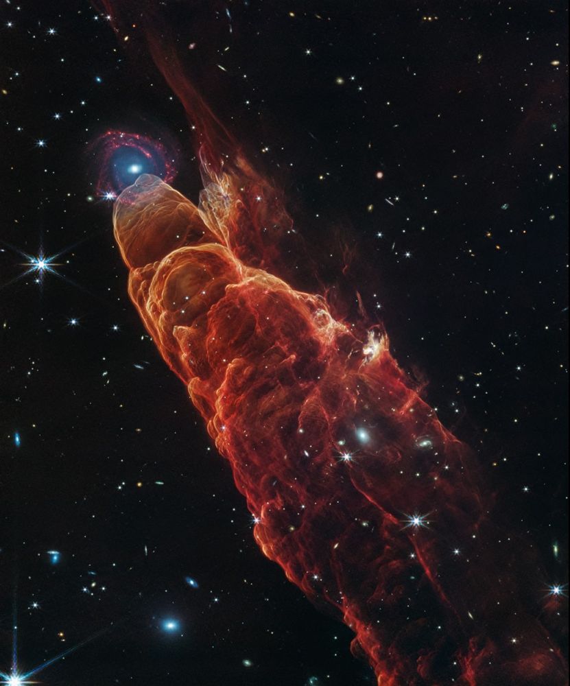 Angled from the upper left corner to the lower right corner is a cone-shaped orange-red cloud known as Herbig-Haro 49/50. This feature takes up about three-fourths of the length of this angle. The upper left end of this feature has a translucent, rounded end. The conical feature widens slightly from the rounded end at the upper right down to the lower right. Along the cone there are additional rounded edges, like edges of a wave, and intricate foamy-like details, as well as a clearer view of the black background of space. In the upper left, overlapping with the rounded end of Herbig-Haro 49/50, is a background spiral galaxy with a concentrated blue center that fades outward to blend with red spiral arms. The background of space is speckled with some white stars and smaller, more numerous, fainter white galaxies throughout.