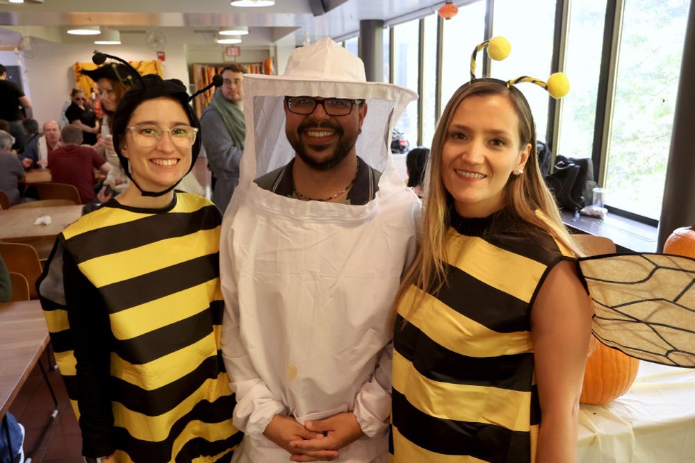 Three people facing the camera, two of them are wearing bee costumes and the one in the middle is dressed as a beekeeper.