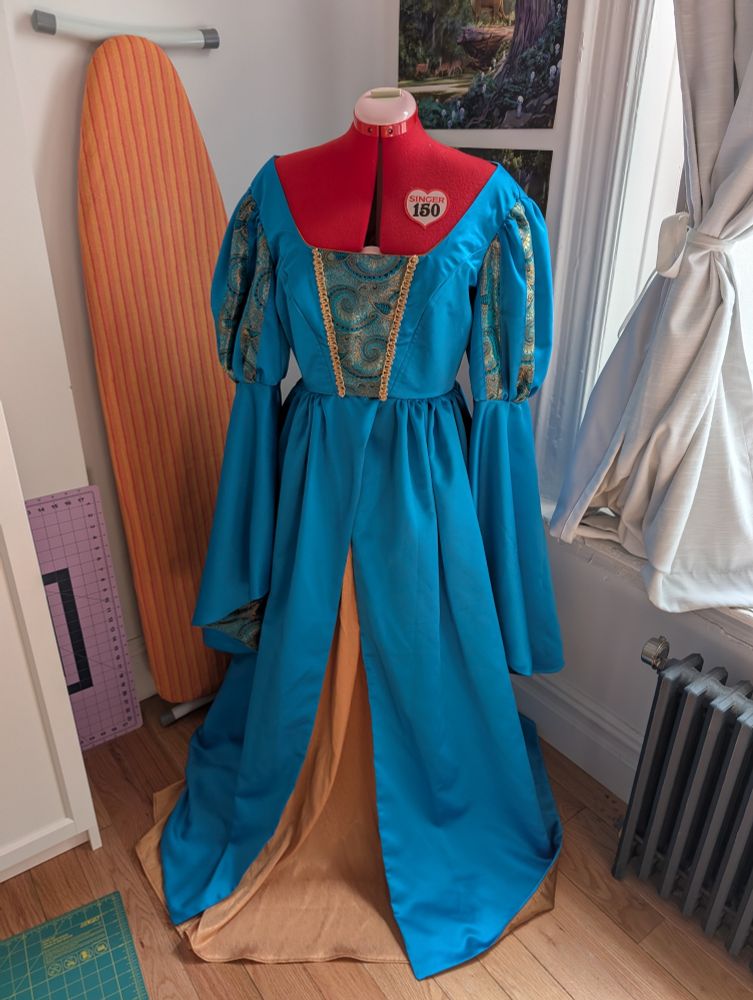 A floor length cerulean dress on a red dress form. It has a split skirt with a gold underskirt beneath, balloon sleeves striped with a paisley blue and gold material and long bell sleeves below that.