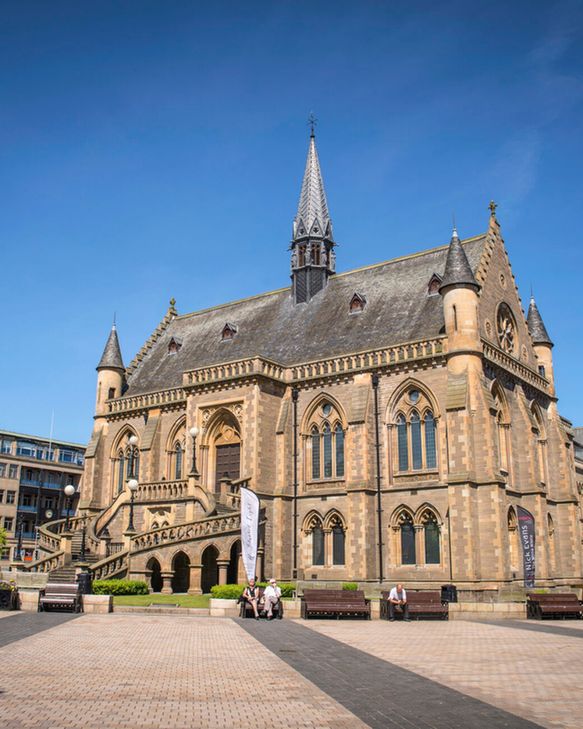 A two-storey building with walls of warm brown stone and a grey tiled roof. It has small turrets and arched windows, and is located in the centre of a small civic square.