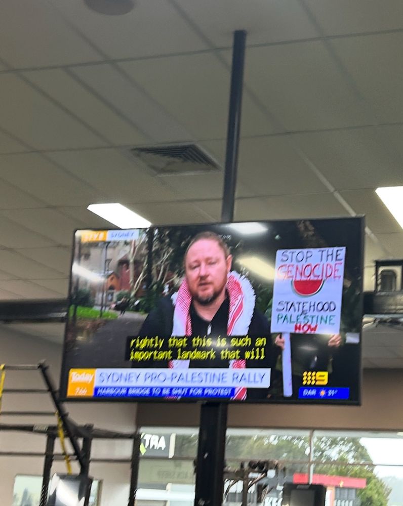Josh Lees is speaking with a kuffiyeh around his neck. Behind him a person is holding up a poster that says ‘Stop the genocide, statehood for Palestine now’ with an image of watermelon on it.