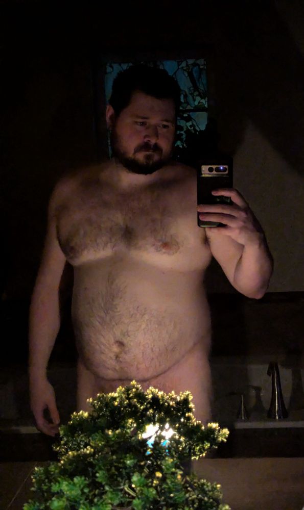 Candlelit nude male taking selfie in front of mirror. A faux bonsai fountain blocks his lower extremities from view