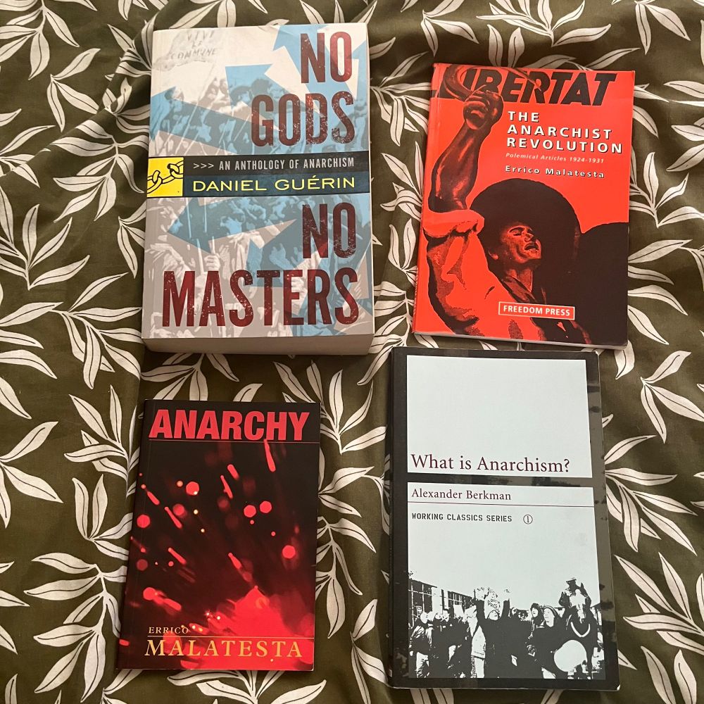 Four books sit on top of a green duvet cover with white olive leaves on it. The top left book is blue and grey, and is “No Gods, No Masters” edited by Daniel Guerin. The top left is red with a black illustration of a peasant with their fist raised, and is “Libertat: The Anarchist Revolution” by Errico Malatesta. The bottom left is black with red splatters and writing, and is “Anarchy”, also by Malatesta. Finally, the bottom left is black and white with a high contrast black photo of a protest across the bottom, and it is “What is Anarchism?” by Alexander Berkman