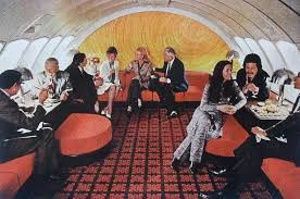 First class lounge on an airplane in the 1960s. Rows of sofas, plush red seats, and a wide carpeted aisle.