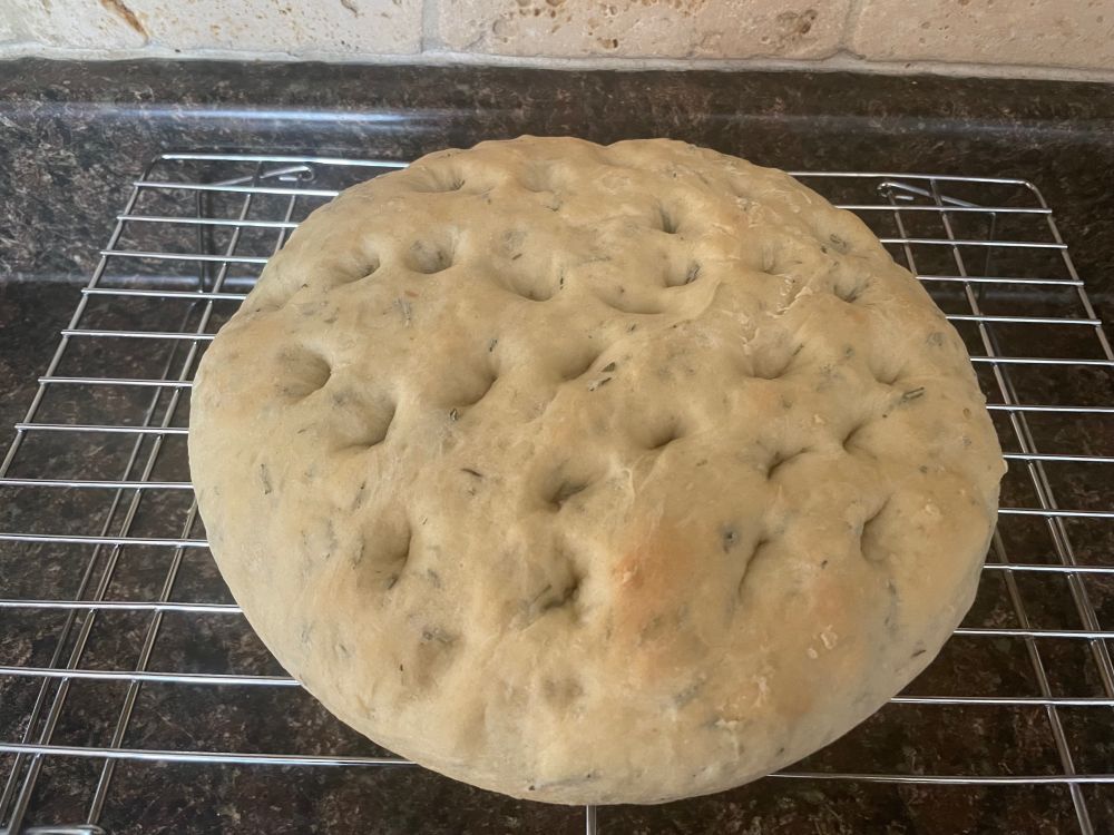 A round loaf on bread with rosemary bits visible and multiple small indentations on the top resting on a wire rack