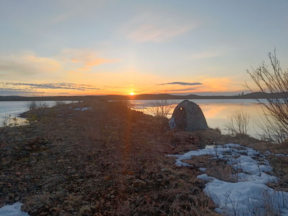 Early morning view by lake. Sun is rising in the middle of the photo. A photo hide is placed by tbe shoreline. Only part of the lake is ice-free. 