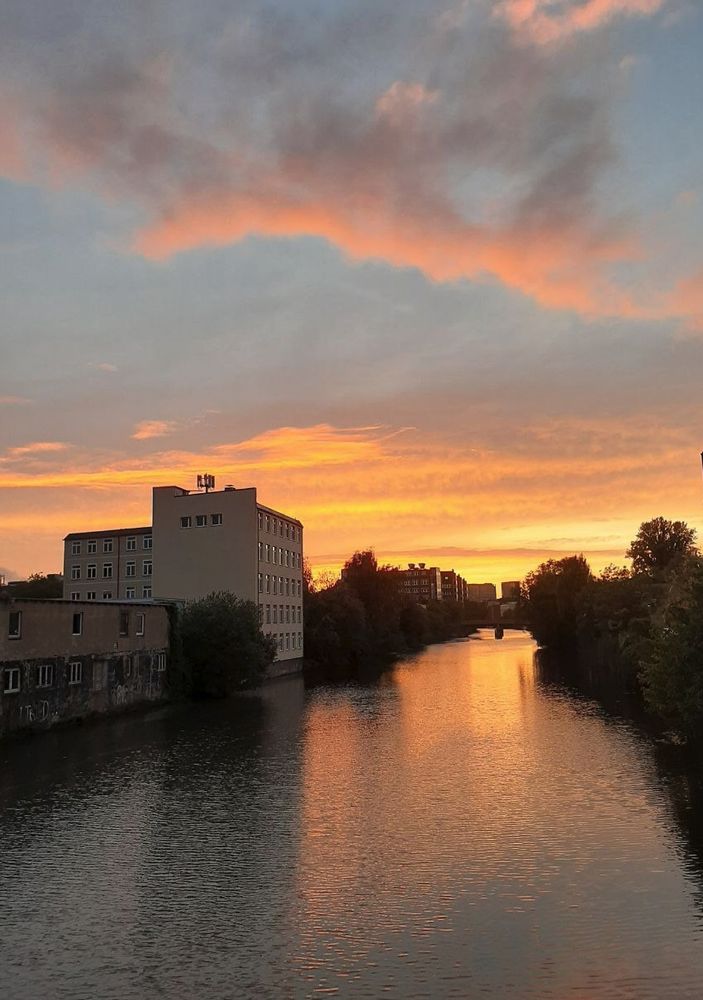 Sonnenuntergang mit sehr orangem Himmel und vielen Wolken über einem Kanal, der Himmel spiegelt sich im Wsser, links und rechts des Kanals Häuser und Büsche.