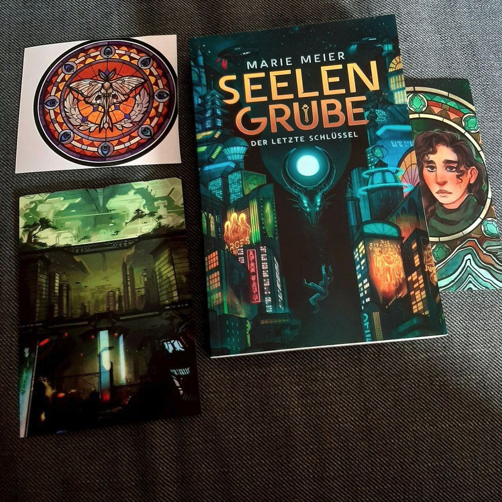 Der Roman "Seelengrube" als Print, dazu ein Sticker, eine Postkarte und ein Lesezeichen mit Motiven aus dem Artwork dazu, leigen auf einer dunkelgrauen Couch.