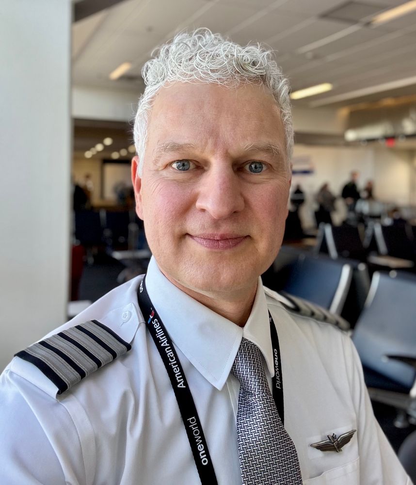 Picture of Eric in an airport wearing a pilots uniform with for stripes on his epaulettes