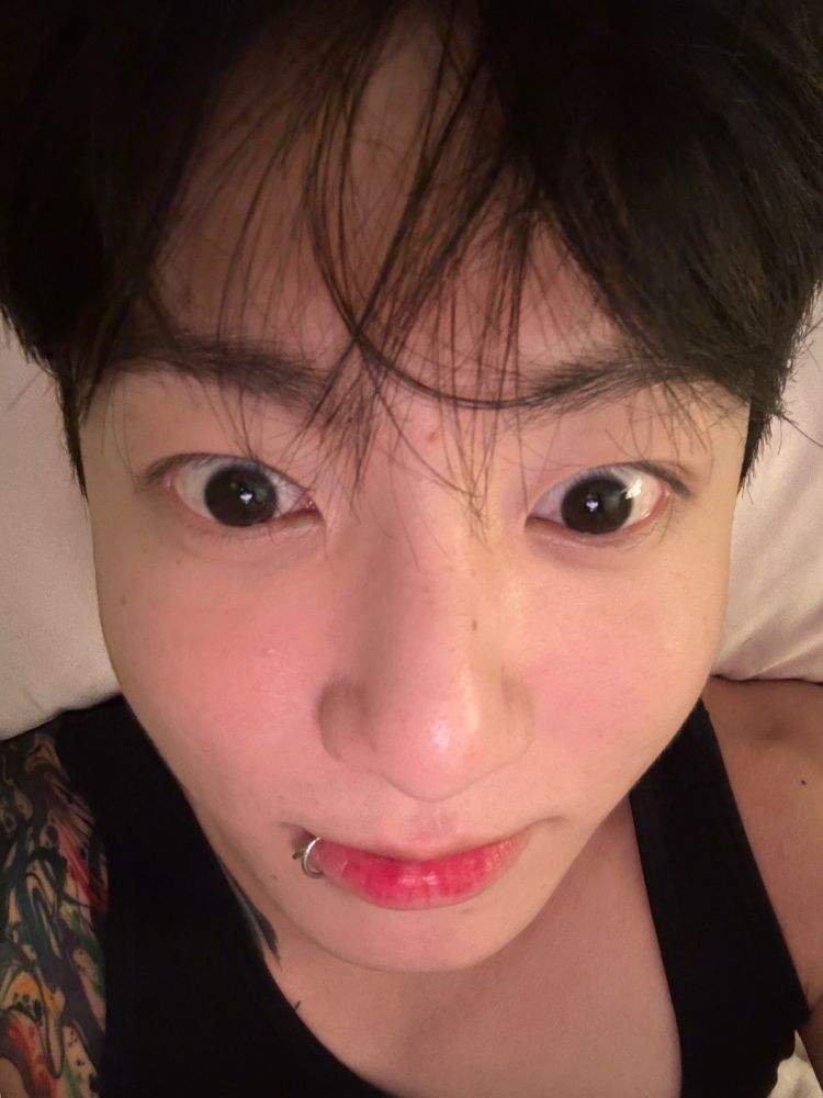 Jungkook selfie, he is in bed his big eyes are looking just below the camera lens and his new shoulder tattoo is peaking under his black tank