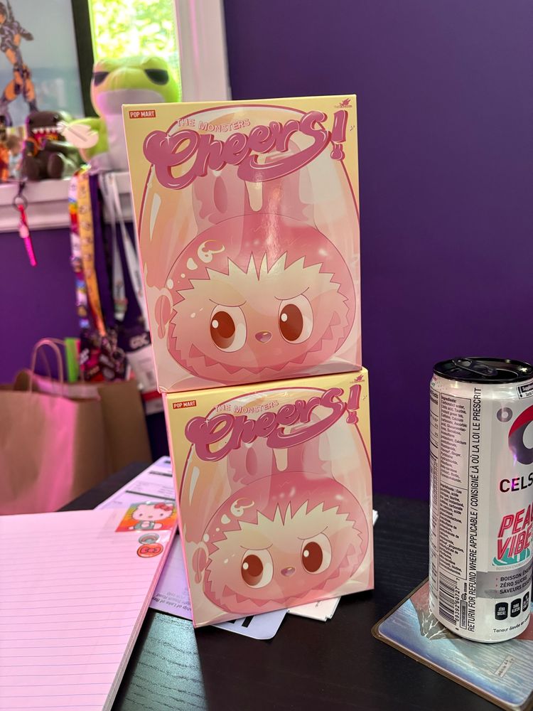 A desktop with two stacked pink-tone boxes that say “Cheers!” in scriptive lettering, with an image showing the box contents to be a drinking tumbler with a grinning, cartoon rabbit-like monster inside the glass. 