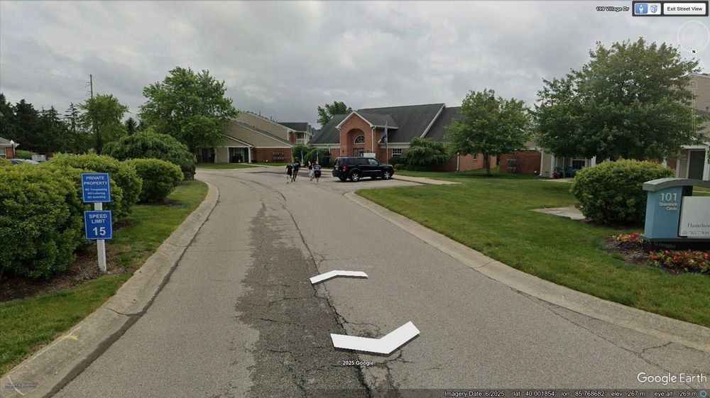 A view down a street. In the short distance, a car sits parked on the right. Several buildings are on the right side of the road. From behind the car, four young people can be seen running towards the camera.