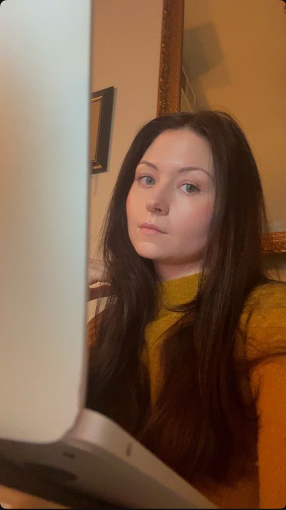 Jamie, brunette with long layers, green eyes, and peachy make up on eyes cheeks and lips, where’s a golden rod, yellow mock turtleneck, and stares down at the camera from behind her laptop as if to say “don’t “