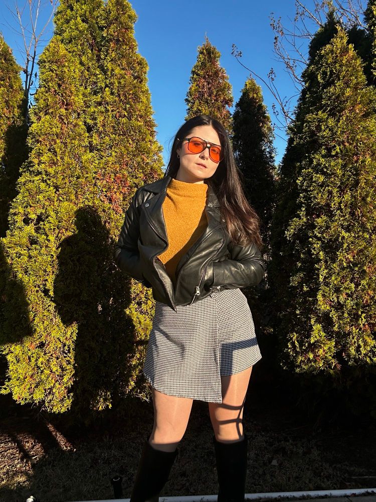 Jamie in a golden sweater, houndstooth skort, and knee high black leather gogo boots 

She now has on a leather jacket from Quince

She has orange tinted lenses in black thick rim aviators that complement her sweater 
