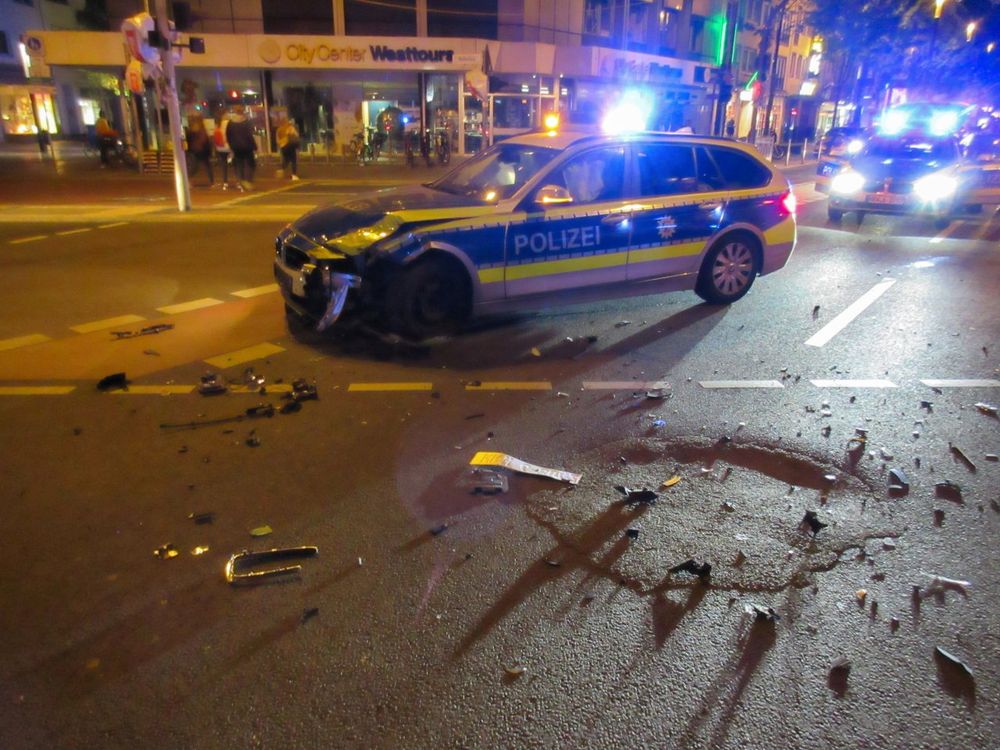 Foto eines Polizeiunfalls am Bertha-von-Suttner-Platz in Bonn in der Nacht vom 1. auf den 2. Mai 2018. Ein Streifenwagen, der durch einen durch die Polizei Bonn verschuldeten Unfall beschädigt wurde, steht gedreht auf einer Kreuzung, dahinter eintreffende Verstärkung. Die Motorhabe des Streifenwagens ist linksseitig stark eingedrückt. Ein Blaulicht, ein gelbes Licht und rotes Rücklicht brennen noch. In dem menschenleeren Streifenwagen sind Airbags aufgeblasen. Auf dem Boden liegen zahlreiche Bruchstücke, die im Scheinwerferlicht glänzen, darunter auch das durch den Unfall abgetrennte und verbogene Nummernschild des Streifenwagens. Eine Gruppe junger Menschen geht im Hintergrund vorbei.

Foto: Marvin Oppong © 2025