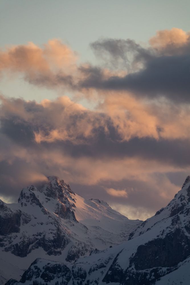 Mountains at sunset with clouds behind them. The mountains and clouds are pink from the sunset.