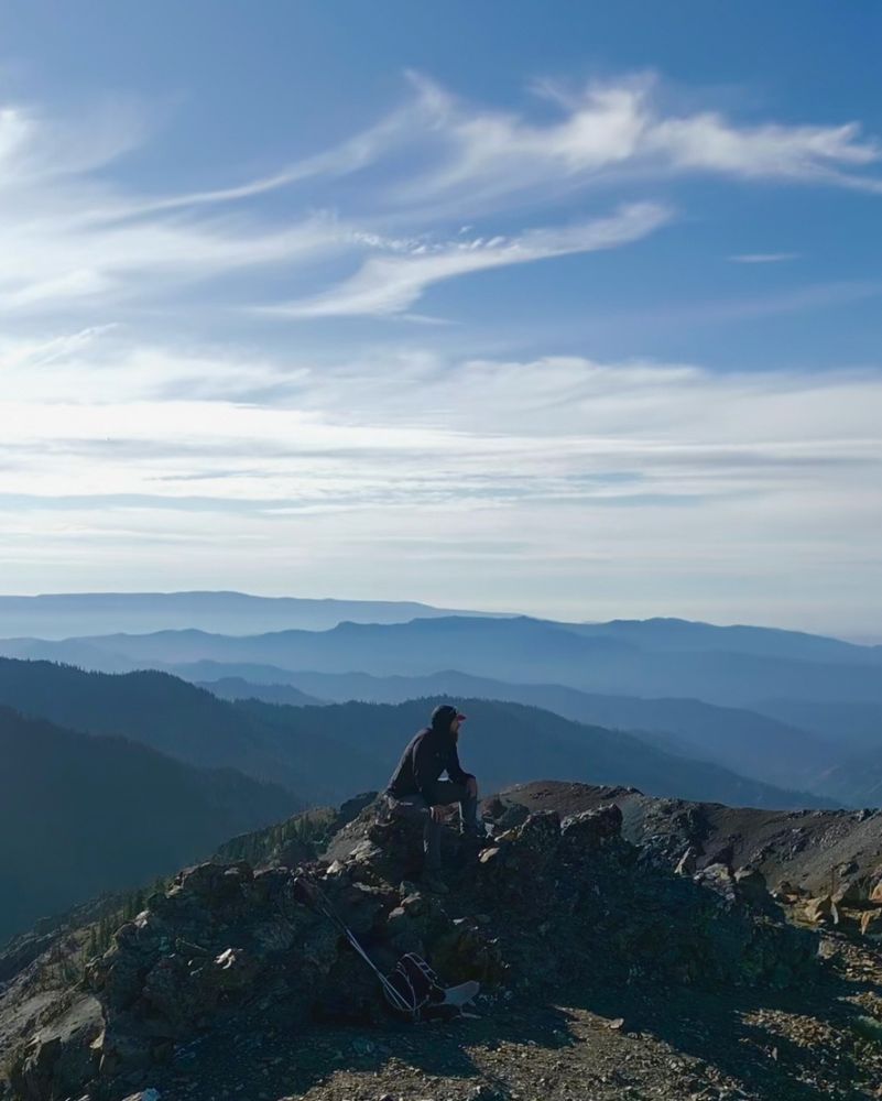 A picture of Tyler sitting on some rocks on top of a mountain. There are layers of mountains in the distance and wispy clouds in the sky.