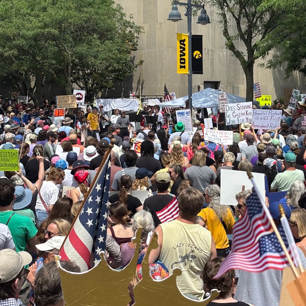 A growing crowd of protestors carrying signs and American flags listen to various speakers make statements critical of the Trump administration. 