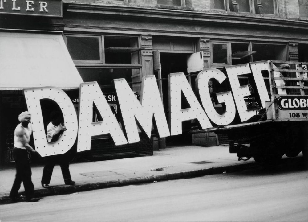 Photo of three workmen loading a truck with a sign made of giant letters reading DAMAGED.